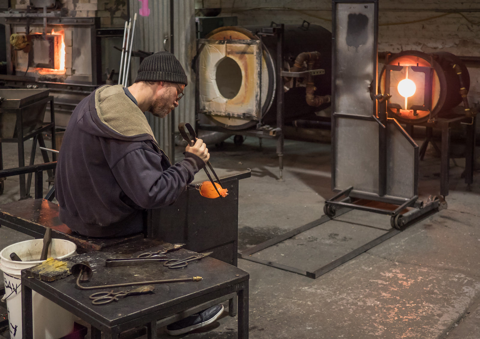 A glassmaker using hand tools to shape glowing glass at the bench
